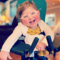 a two year old girl with glasses smiles, she is supported by a stander